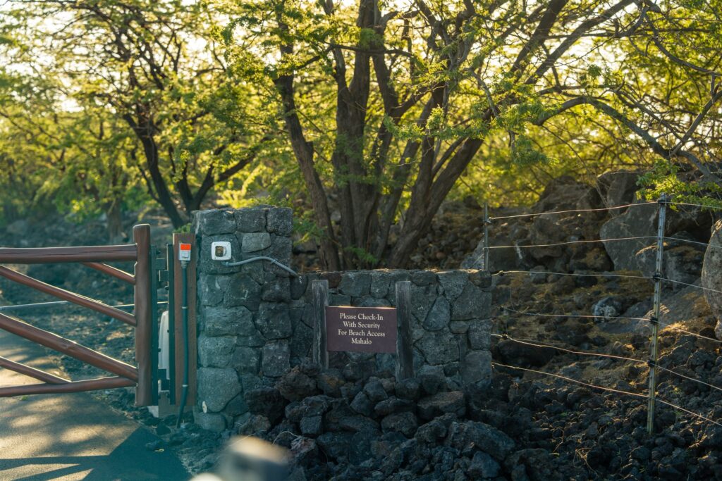 A stone wall and metal gate block a path at Kikaua Point Park, surrounded by trees and rocks. A sign on the wall reads, Please Check-In With Security for Beach Access Mahalo. Sunlight filters through the branches.