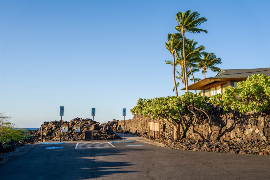 A small paved parking lot with reserved accessible parking spaces at Kikaua Point Park overlooks rocky lava terrain, green shrubs, and tall palm trees next to a building under a clear blue sky.