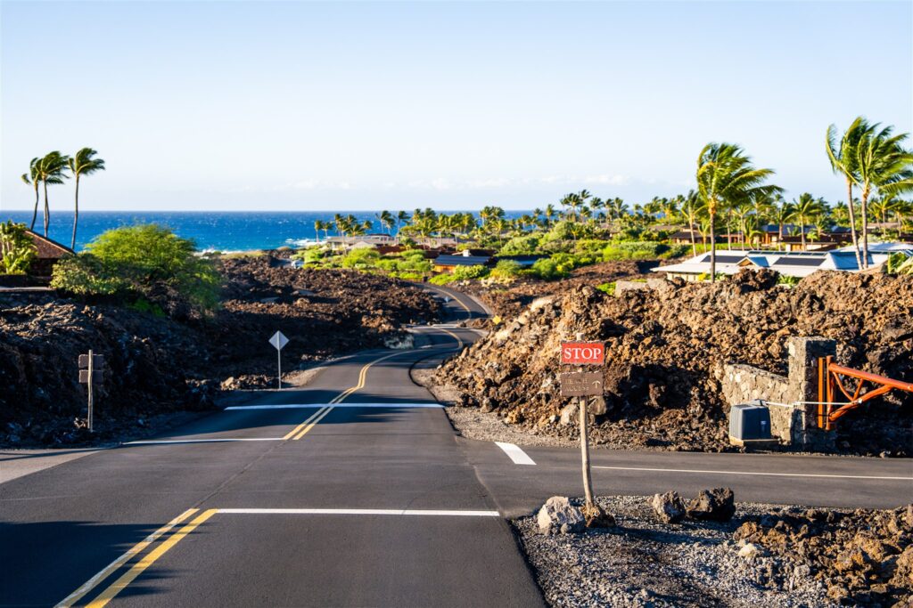 A paved road winds through dark volcanic rocks at Kikaua Point Park, lined with palm trees and tropical vegetation, leading towards blue ocean under a clear sky. A small red stop sign is visible on the right side of the road.