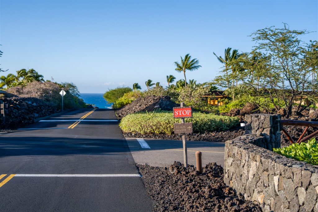A paved road lined with stone walls and tropical plants leads toward the ocean at Kikaua Point Park under a clear blue sky. A small stop sign and lava rocks are visible at an intersection in the foreground.