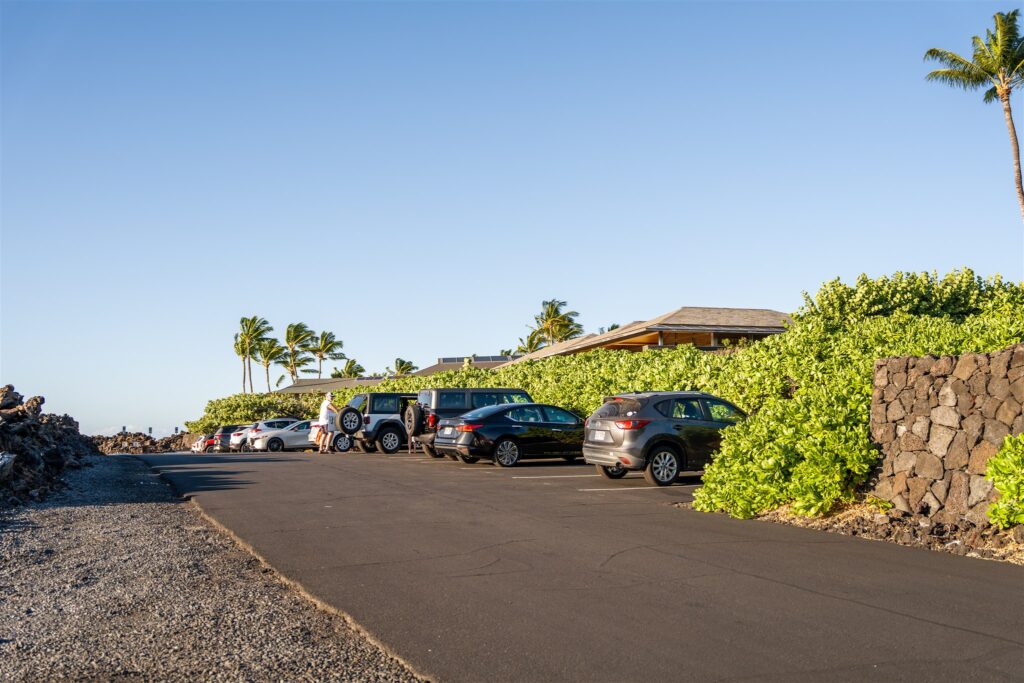 A small parking lot at Kikaua Point Park with several cars parked along a curb, surrounded by lush green bushes and palm trees under a clear blue sky. A few people are near the cars, and a stone wall lines one side.