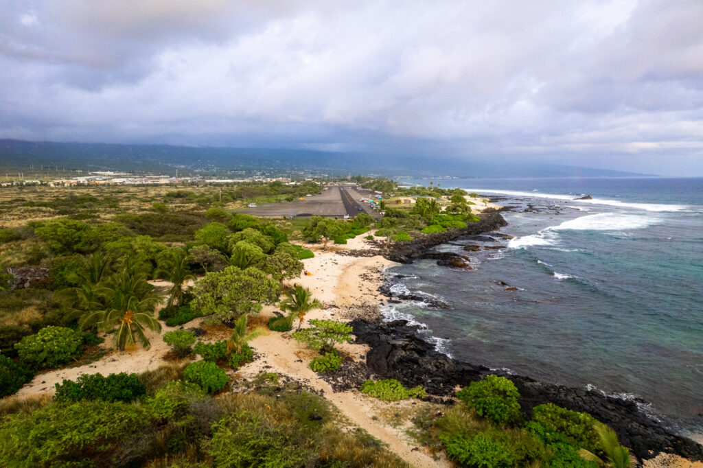 Aerial view of a tropical coastline with sandy beach, black volcanic rocks, lush green trees, and a small airport runway near the ocean under a cloudy sky.