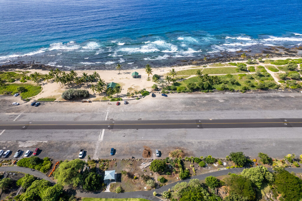 Aerial view of a coastal road and parking lot beside a sandy beach with palm trees, green shrubs, and blue ocean waves crashing against a rocky shoreline. Several cars are parked near the beach and along the roadside.