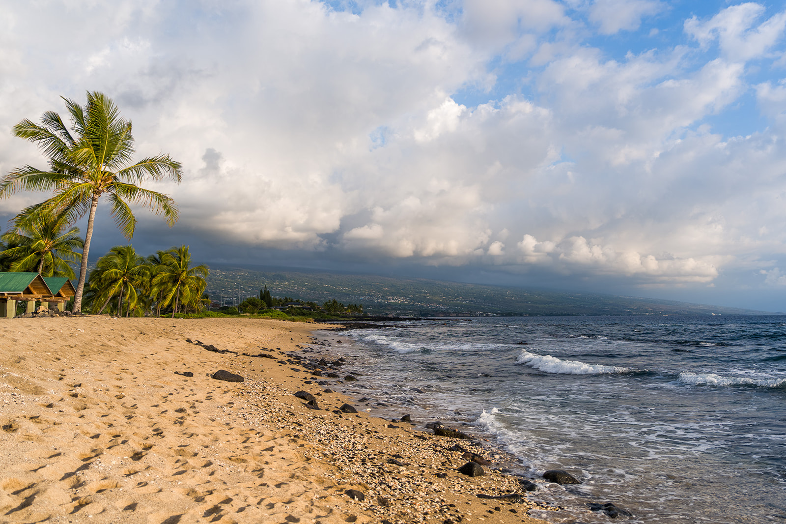 A sandy beach with scattered rocks and palm trees along the shore, gentle waves washing up, and a mountainous coastline in the distance under a partly cloudy sky, of Old Kona Airport Beach.