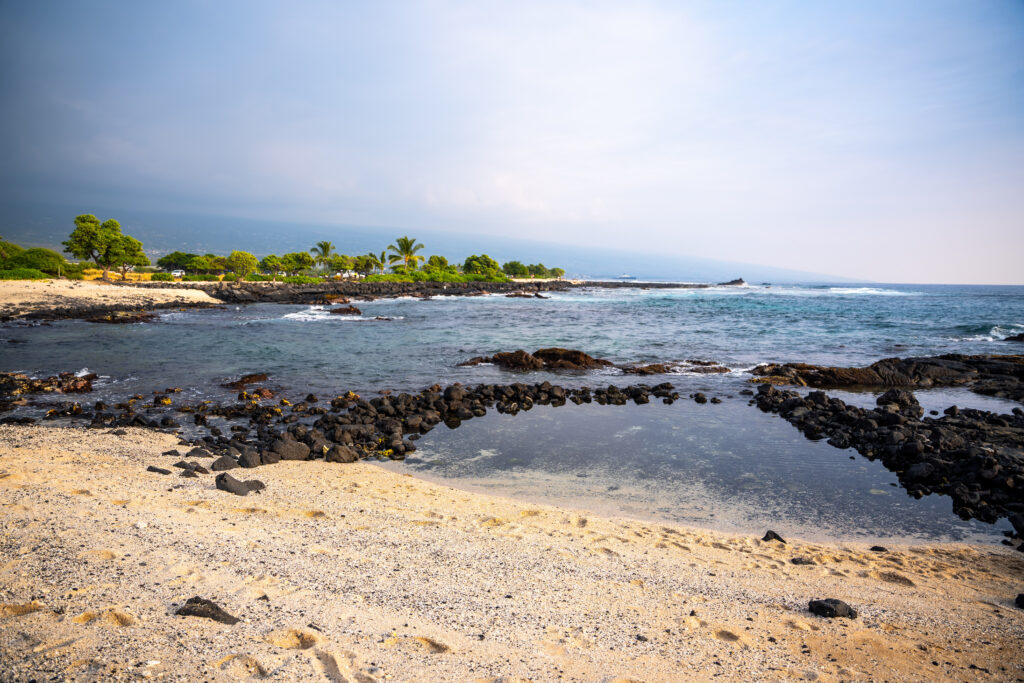 Sandy beach with black volcanic rocks, calm tidal pool, blue ocean waves, and green trees along the shoreline under a slightly cloudy sky.