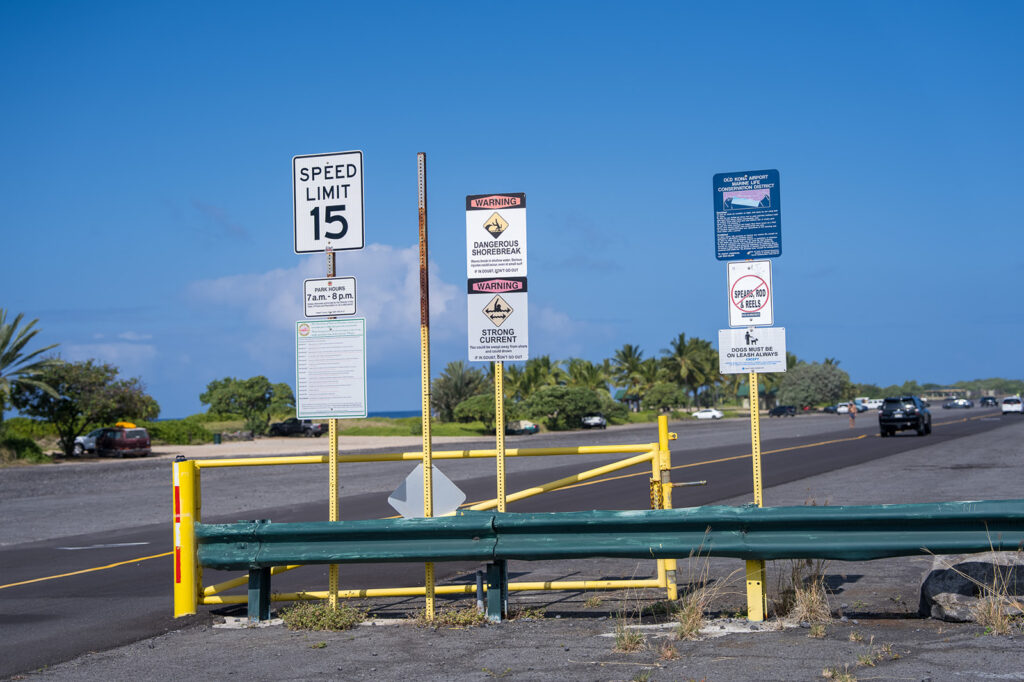A row of warning and informational signs, including a speed limit sign, stands near a yellow gate and roadside barrier along a sunny coastal road with palm trees and cars in the background.