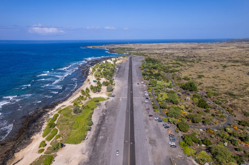 Aerial view of a long, narrow road running parallel to the coastline with blue ocean waves on one side and dry, grassy land with scattered trees and parked cars on the other under a clear sky.