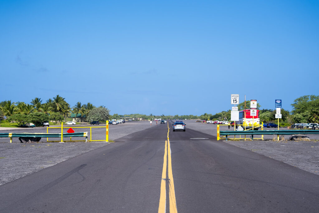 A paved road with double yellow lines leads to a parking lot surrounded by greenery and palm trees under a clear blue sky. There are cars parked, signs, and barriers along the road.