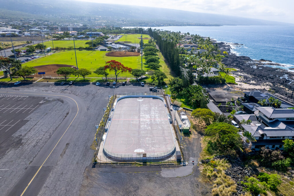 Aerial view of a coastal recreation area with an outdoor sports rink, parking lot, baseball fields, trees, nearby buildings, and the ocean to the right. The shoreline features rocky terrain and waves.
