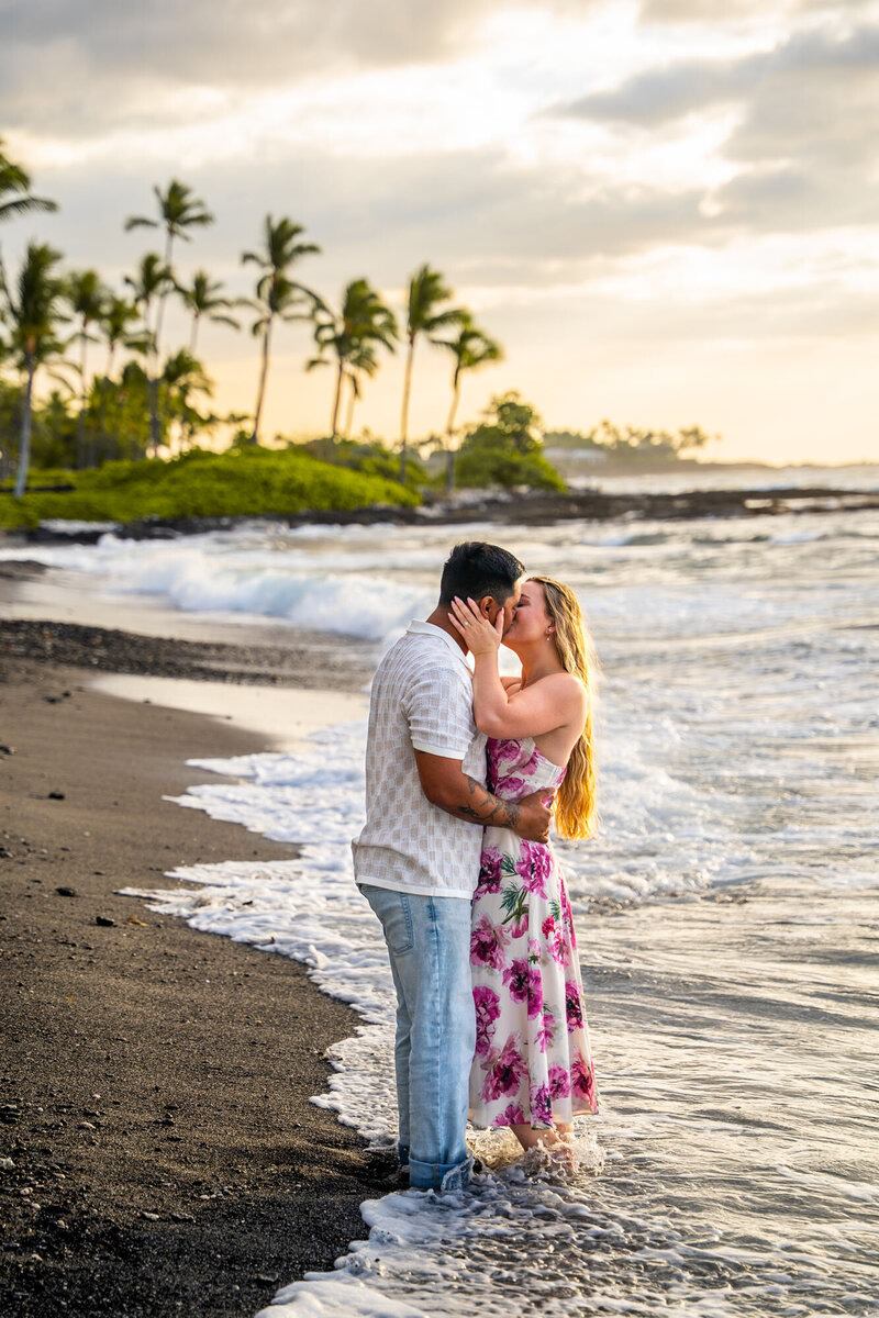 Couple kissing on the beach with palm trees in the background.