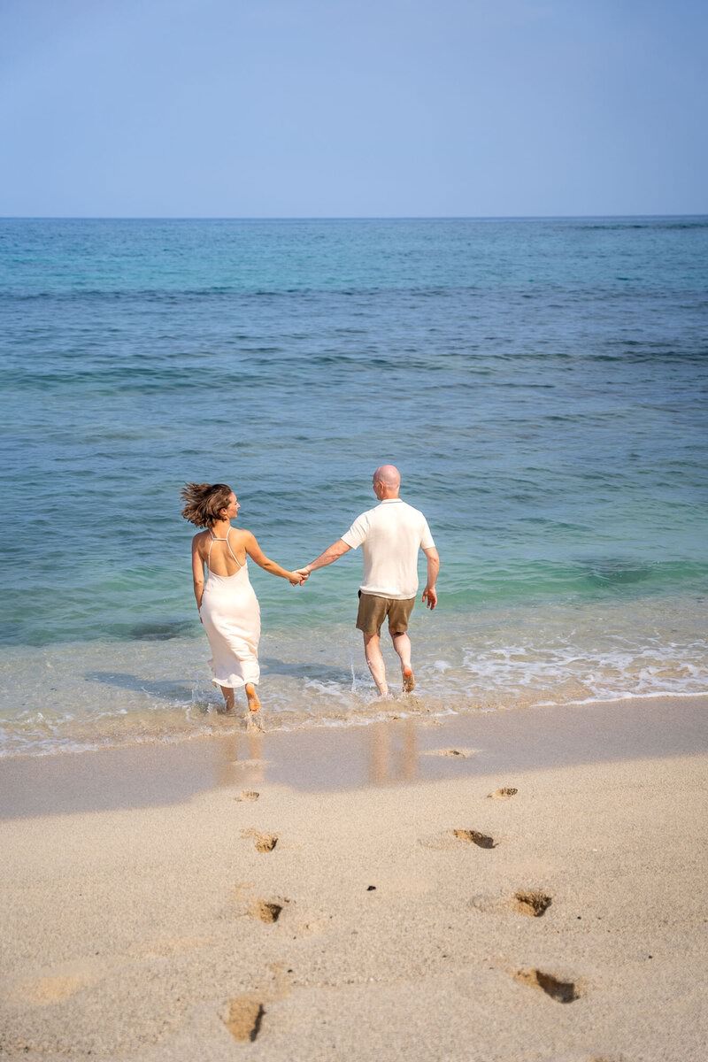 During engagement photos, a couple runs into the ocean together holding hands on a Hawaii beach.
